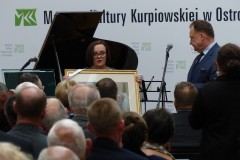 Kobieta stoi pokazując obraz stojąc obok mikrofonu wraz z mężczyzna przed widownią na tle napis muzeum kultury kurpiowskiej i fortepian