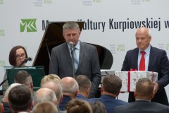 Mężczyzna stoi przy mikrofonie przed widownią na tle napisu muzeum kultury kurpiowskiej i fortepianie