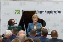 Kobieta stoi przy mikrofonie przed widownią na tle napisu muzeum kultury kurpiowskiej i fortepianie