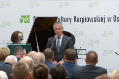 Mężczyzna stoi przy mikrofonie przed widownią na tle napisu muzeum kultury kurpiowskiej i fortepianie