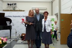 Trzy osoby pozujące dla zdjęcia na tle napisu Muzeum Kultury Kurpiowskiej i fortepianie
