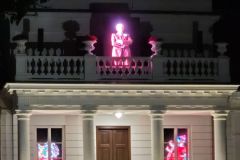 Kilka osób przed białym budynkiem, na balkonie iluminacja przedstawiająca postać mężczyzny stojącegoi z założonymi rękami