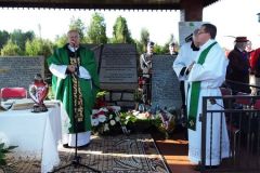 Pod zadaszeniem w plenerze stoi dwóch księży i mężczyzna w komży, z lewej strony fragment stoły z białym obrusem. W tle kamienne tablice z napisami, dalej dwóch mężczyzn w mundurach, z prawej w tle inne osoby.