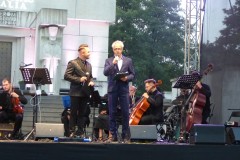 Dwóch mężczyzn stojąc na scenie zapowiada koncert, w tle grupa muzyków gra.