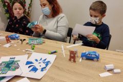 Kobieta oraz dwoje dzieci siedząc przy stole wykonują zabawki z papieru.