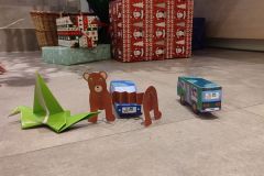 Zabawki z tektury wykonane przez uczestników rodzinnych zajęć twórczych. W tle fragment choinki bożonarodzeniowej, pod którą leżą prezenty.