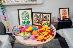 Na okrągłym stoliczku leżą ozdoby choinkowe, wykonane z kolorowego papieru, za nimi stoją obrazy z wycinankami kurpiowskimi.