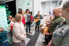 Kilka osób stoi, dwie z nich grają na instrumentach, przed nimj stoi dwójka harcerzy jeden z nich trzyma latarenkę.