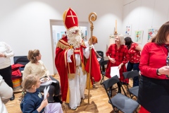 Święty Mikołaj w przebraniu biskupa, dwie dziewczynki. Kilka kobiet w czerwonych ubraniach.