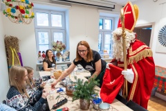 Przy jednym stole siedzą dwie dziewczynki, dwie kobiety i Święty Mikołaj w przebraniu biskupa. W tle są dwa duże okna, a w lewym roku snopek zboża.