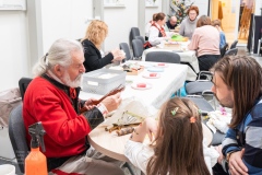 Mężczyzna w stroju kurpiowskim pokazuje dziewczynce i dorosłemu mężczyźnie wiklinowe gałązki. W tle robione są bombki z opłatka.