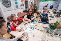 Przy jednym stole siedzi kilkoro dzieci i trzy kobiety, ozdabiają szklane słoiki.