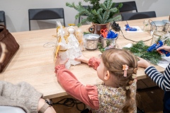 Mała dziewczynka trzymająca dwa aniołki ze styropianu, po jej prawej dziecko przyklejające gałązki choinki do kartki.