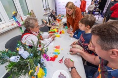 Przy jednym stole siedzą trzy osoby starsze, jedna pani jest w stroju kurpiowskim, pokazuje trzem chłopcom jak wykonać aniołki z bibułki.