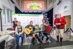 Pięć osób śpiewających, jedna kobieta grająca na gitarze, mężczyzna grający na akordeonie obok niego stoi mężczyzna ubrany w strój kurpiowski.