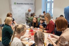Dzieci, młodzież i dorośli siedząc lub stojąc przy stołach wykonują prace plastyczne. Przy dalszym stole siedzą harcerze. W tle okno oraz ścianka z nazwą Muzeum i logami
