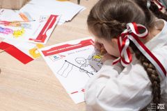 Dziewczynka z biało-czerwoną wstążką we włosach siedząc przy stole koloruje obrazek