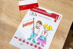 Kolorowanka przedstawiająca chłopczyka i dziewczynkę, chłopiec trzyma biało-czerwoną flagę. Obok kolorowanki leży mała papierowa biało-czerwona flaga.