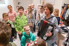 W pomieszczeniu stoi mężczyzna grając na akordeonie, a wokół niego kilkanaście osób – dorośli i dzieci.