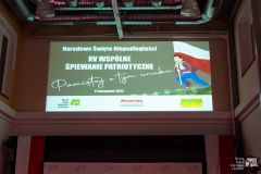 Banner z napisem Narodowe Święto Niepodległości XV wspólne śpiewanie patriotyczne Pamiętaj o tym wnuku oraz ilustracją chłopca biegnącego z biało-czerwoną flagą wyświetlony na ścianie