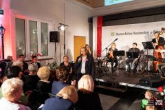 Na muzealnym dziedzińcu kobieta przemawia przez mikrofon do publiczności. W tle muzycy na scenie.