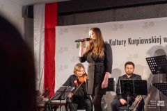 Kobieta stojąc na scenie śpiewa. W tle siedzą muzycy.