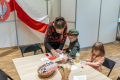 Przy stole siedzi dziewczynka, obok stoi chłopiec i kobieta. Na stole biało-czerwone elementy do wykonania papierowej flagi. W tle na stelażu białoczerwona flaga oraz fragment dawnego godła.