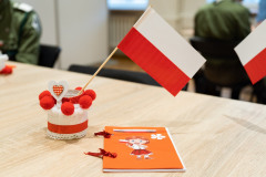 Na stole stoi mały lampionik zdobiony biało-czerwonymi elementami, a w nim wstawiona biało-czerwona flaga wykonana z papieru. Obok leży zdobiony, papierowy notesik z wizerunkiem dziewczynki trzymającej polską flagę.