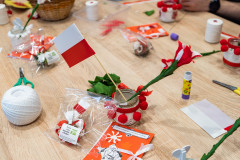 Na stole stoi mały lampionik, a w nim wstawiona papierowa flaga biało-czerwona oraz kwiatek z bibuły. Wokół leży wiele materiałów oraz przyborów do tworzenia, nożyczki, kleje biurowe, nici, skrawki bibuły.