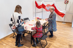 Przy stole siedzi dziewczynka oraz chłopiec, obok stoją dwie kobiety. Na stole elementy do tworzenia biało-czerwonych flag, materiały edukacyjne, kilka papierowych flag w drewnianym pojemniczku, pleciony koszyczek, małe lampioniki, w jednym lampioniku kwiatek z bibuły. W tle na stelażu rozpostarta biało-czerwona flaga, oraz dawne godło.