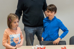 Dziewczynka i chłopiec, pomiędzy nimi mężczyzna, stoją za stołem, na którym są trzy małe lampioniki zdobione biało-czerwonymi elementami oraz materiały i przybory to tworzenia.