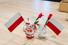 Fragment stołu, na stole dwa małe lampioniki zdobione biało-czerwonymi elementami,  w lampionikach w każdym lampioniku papierowa flagi oraz  kwiat z bibuły.