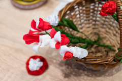 Fragment plecionego koszyczka stojącego na stole widziany od góry. W koszyczku kilka a biało-czerwonych kwiatków z bibuły. Obok koszyczka mała kokarda narodowa.