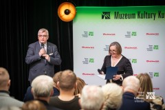 Dwie osoby z mikrofonami w dłoniach stoją przed publicznością, w tle ścianka z logami MKK i marki Mazowsze
