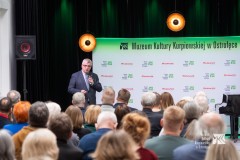 Prelegent z mikrofonem w dłoni stoi przed publicznością, w tle ścianka z logami MKK i marki Mazowsze
