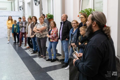 W pomieszczeniu kilkanaście osób, dorośli i troje dzieci stoją w szeregu, słuchając przemówienia autora wystawy. W tle ściana z oknami, w oddali fragment napisu Muzeum Kultury Kurpiowskiej w Ostrołęce.