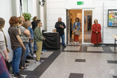 Kilkanaście osób zebranych na wernisażu, słucha przemówienia autora wystawy. W tle drzwi, przed którymi stoją dwie kobiety, z jedną z kobiet stoi mała dziewczynka.