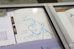 Postać z komiksu w gablocie wystawowej. W chmurce napis DLA STASZKA. Obok postaci podpis autora Sławek Kiełbus.