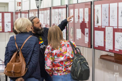 W pomieszczeniu, mężczyzna oraz dwie kobiety stoją przy rysunkach eksponowanych na wystawie. Mężczyzna wskazuje na jeden z rysunków.