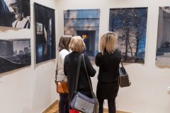 Trzy kobiety przyglądają się fotografiom wiszącym na ścianie sali wystawowej.