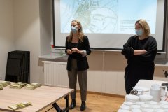 Dwie kobiety stoją na tle ekranu z wyświetlaną prezentacją. Z lewej strony, na stole leżą  publikacje. Z prawej strony, na stole elementy serwisu kawowego.