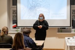 Kobieta w maseczce stoi na tle ekranu z wyświetloną prezentacją. Za ekranem fragmenty okien. Z lewej strony ekranu, na parapecie okna we flakonie stoi papierowa biało-czerwona flaga. Z lewej strony przy stołach siedzą trzy osoby zwrócone w stronę stojącej kobiety.