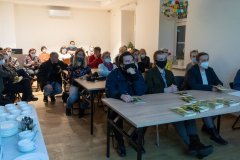 Przy stołach siedzi wiele osób zwróconych w stronę ekranu z prezentacją znajdującego się poza kadrem. Z lewej strony na stole fragment serwisu kawowego.