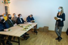 Z lewej strony sześć osób w maseczkach siedzi przy stole na którym leżą publikacje. Zwróceni w prawą stronę kadru, gdzie stoi kobieta w maseczce trzymająca w dłoni egzemplarz przewodnika. Stojąca kobieta zwrócona w stronę siedzących osób.