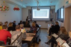 Przy stołach siedzą ludzie zwróceni w stronę ekranu z wyświetloną prezentacją. Niektórzy trzymają w dłoniach otwarte egzemplarze przewodnika. Z lewej strony ekranu stoi kobieta w maseczce prezentująca egzemplarz przewodnika. Za ekranem  fragmenty okien. Z lewej strony, pod sufitem wiszą kierce.