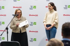 Dwie kobiety stoją przed widownią, jedna z nich przemawia do mikrofonu. W tle ścianka z logami marki Mazowsze oraz  MKK
