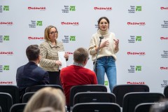 Dwie kobiety stoją przed widownią, jedna z nich przemawia do mikrofonu. W tle ścianka z logami marki Mazowsze oraz  MKK