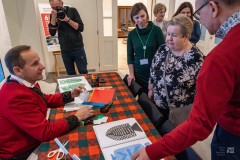 Sześć osób stoi wokół stołu, jeden mężczyzna fotografuje. Przy stole siedzi mężczyzna w stroju kurpiowskim, na stole leżą wycinanki, nożyce, papier kolorowy