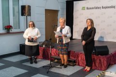 Trzy kobiety stoją obok siebie na muzealnym, krytym dziedzińcu. Kobieta stojąca w środku trzyma w dłoniach kartkę i przemawia do mikrofonu.