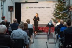 Kobieta stoi przy mikrofonie na muzealnym, krytym dziedzińcu, trzyma w dłoniach folder. Przed nią widownia. W przejściu między rzędami krzeseł stoi statyw z rejestrującym spotkanie telefonem komórkowym.  W tle ścianka z logotypami Muzeum Kultury Kurpiowskiej, z prawej strony kilkumetrowa choinka bożonarodzeniowa.